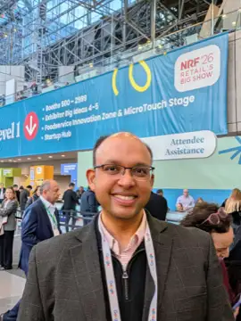 A man poses for a photo at NRF '26 Retail's Big Show, in front of a banner listing exhibitor booths and a Google lanyard.