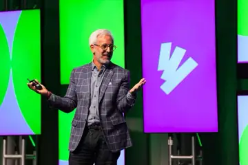 A man speaks on stage gesturing with hands, with green and purple screen backdrops behind him.